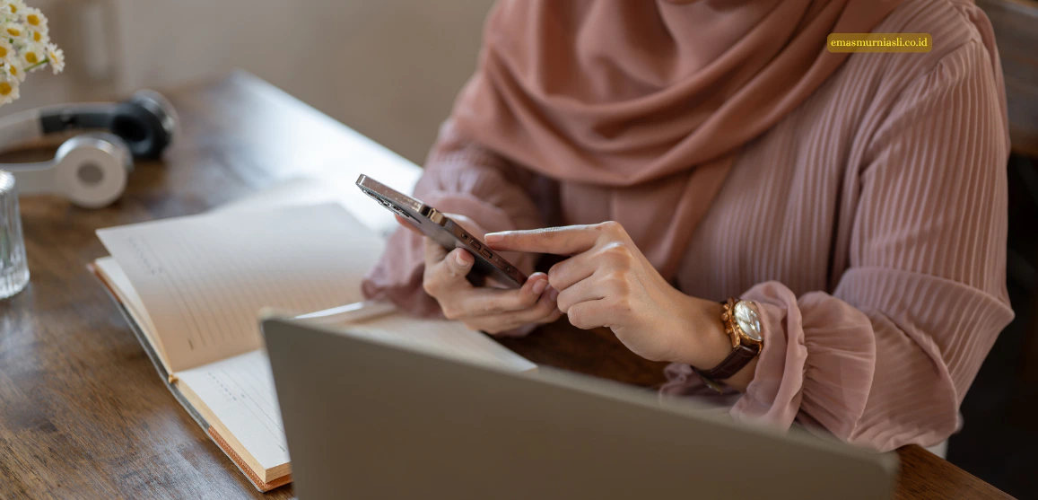 Tangan seorang wanita berhijab yang sedang menggunakan smartphone di meja dengan laptop dan buku catatan.   
