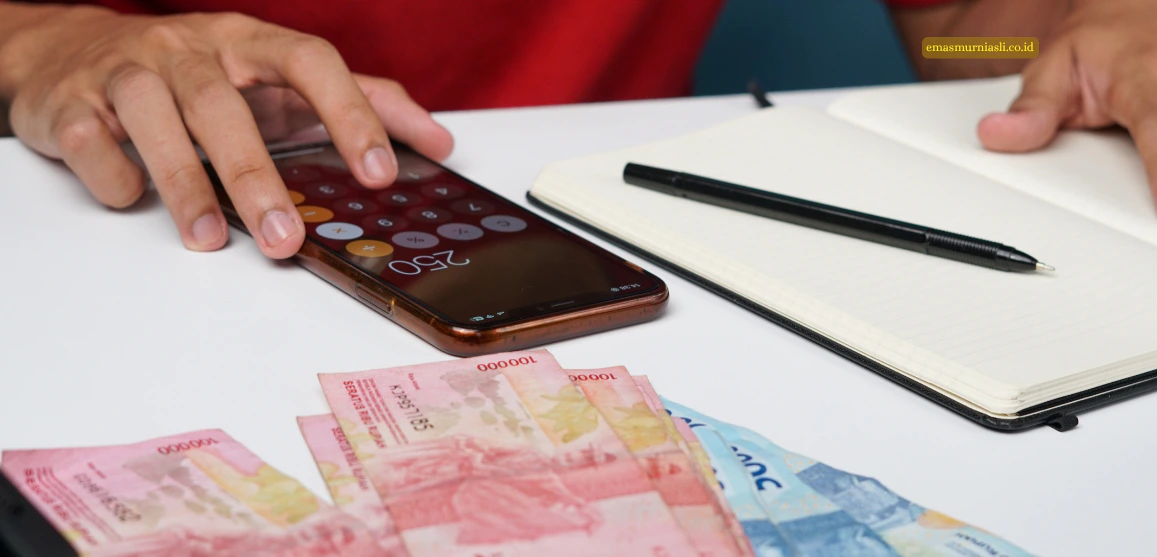 Tangan sedang menghitung keuangan menggunakan kalkulator ponsel di samping lembaran uang Rupiah dan buku catatan.